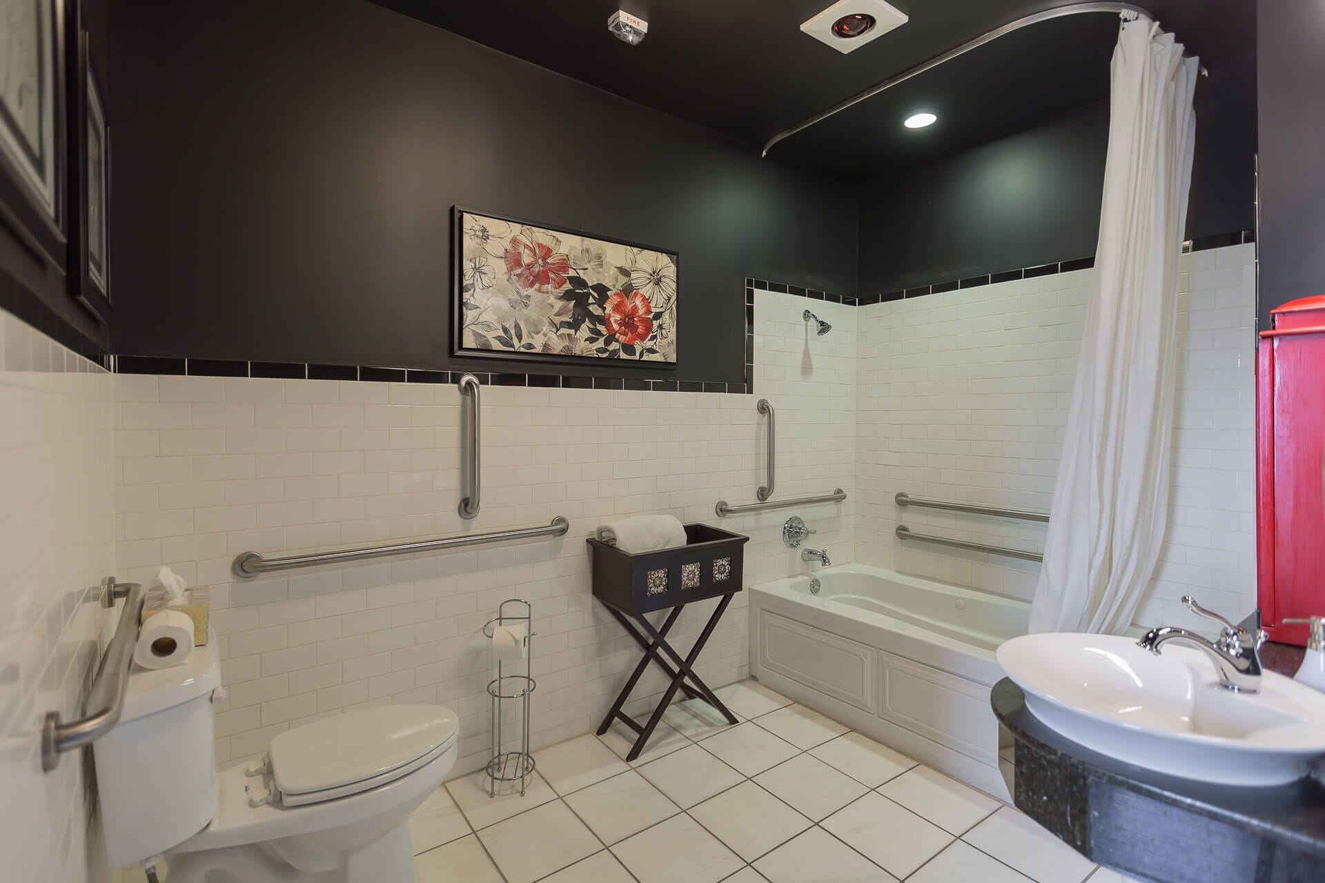 Un baño moderno con azulejos blancos, un inodoro, un lavamanos y una bañera. Hay barras de apoyo y una cortina de ducha.