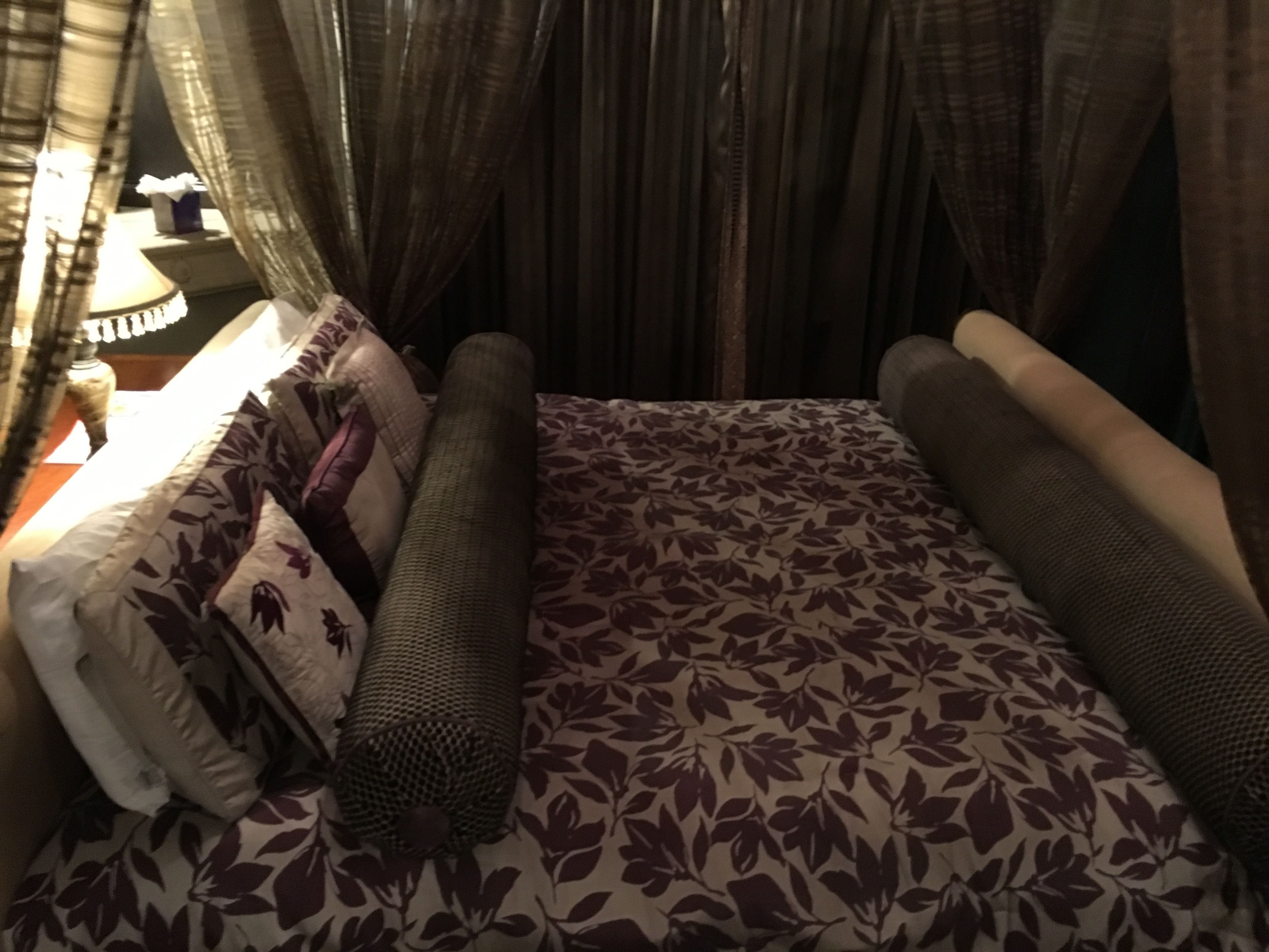 Una cama con colcha estampada de flores, varios cojines y cabecera con cortinas.