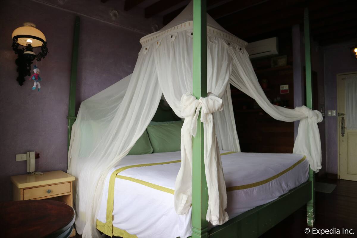 Uma cama de dossel com quatro colunas verdes e cortinas brancas, uma mesinha de cabeceira com abajur e um telefone fixado na parede.