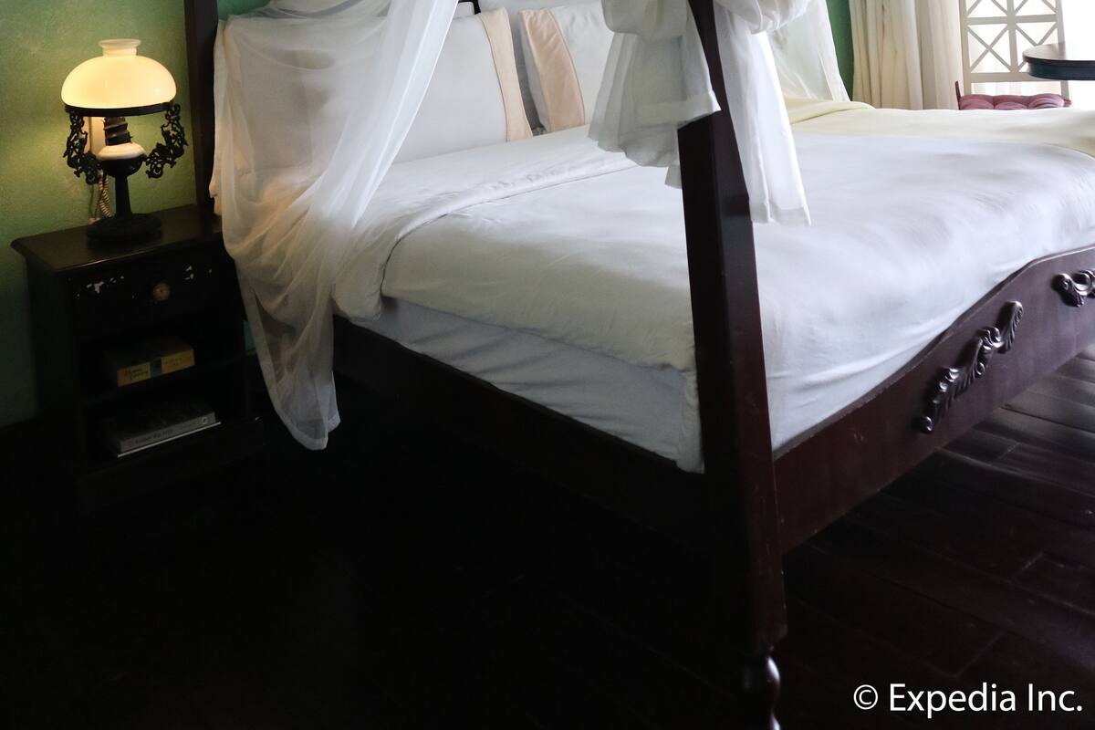 Uma cama de dossel com roupa de cama branca e um toldo, uma mesa de cabeceira com uma luminária e um teto de madeira.