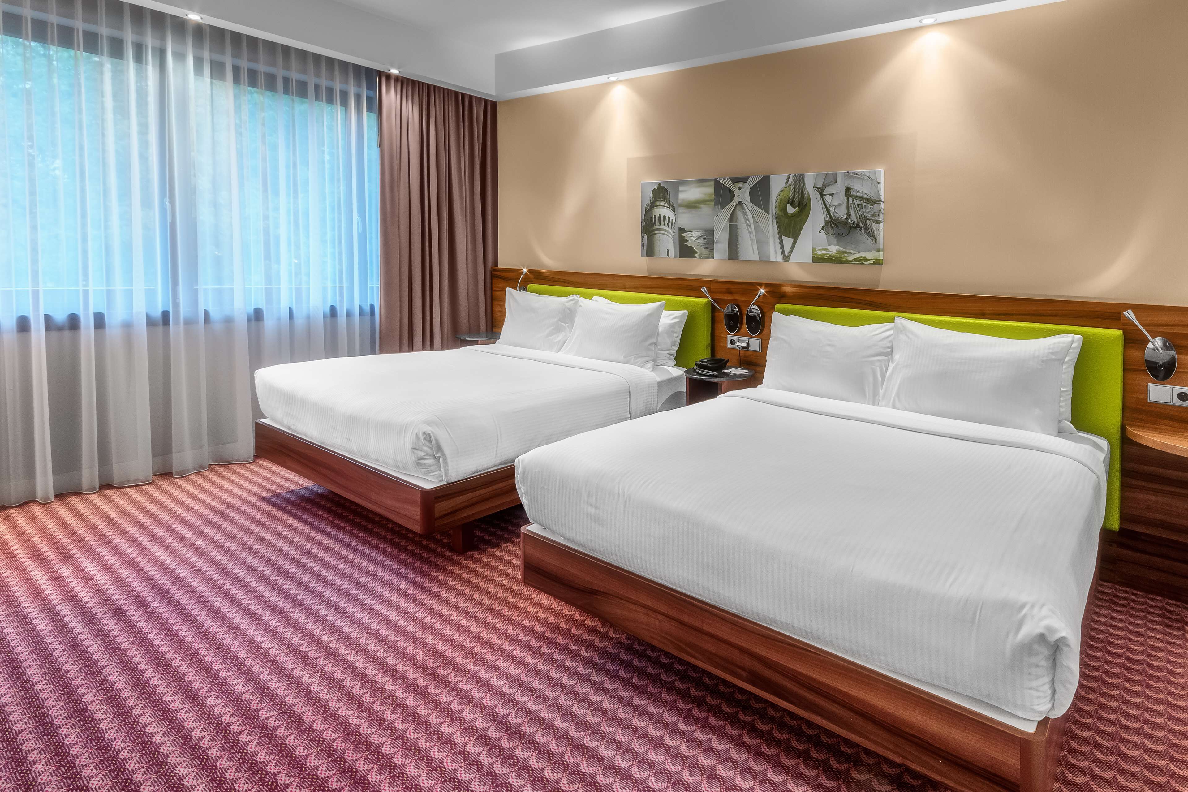A hotel room with two beds, a large window with curtains, a wooden headboard, and a wall-mounted artwork.