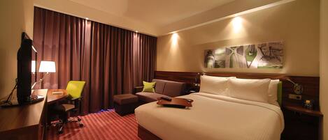A hotel room with a large bed, a desk with a computer, a chair, a sofa, and a wall-mounted TV.