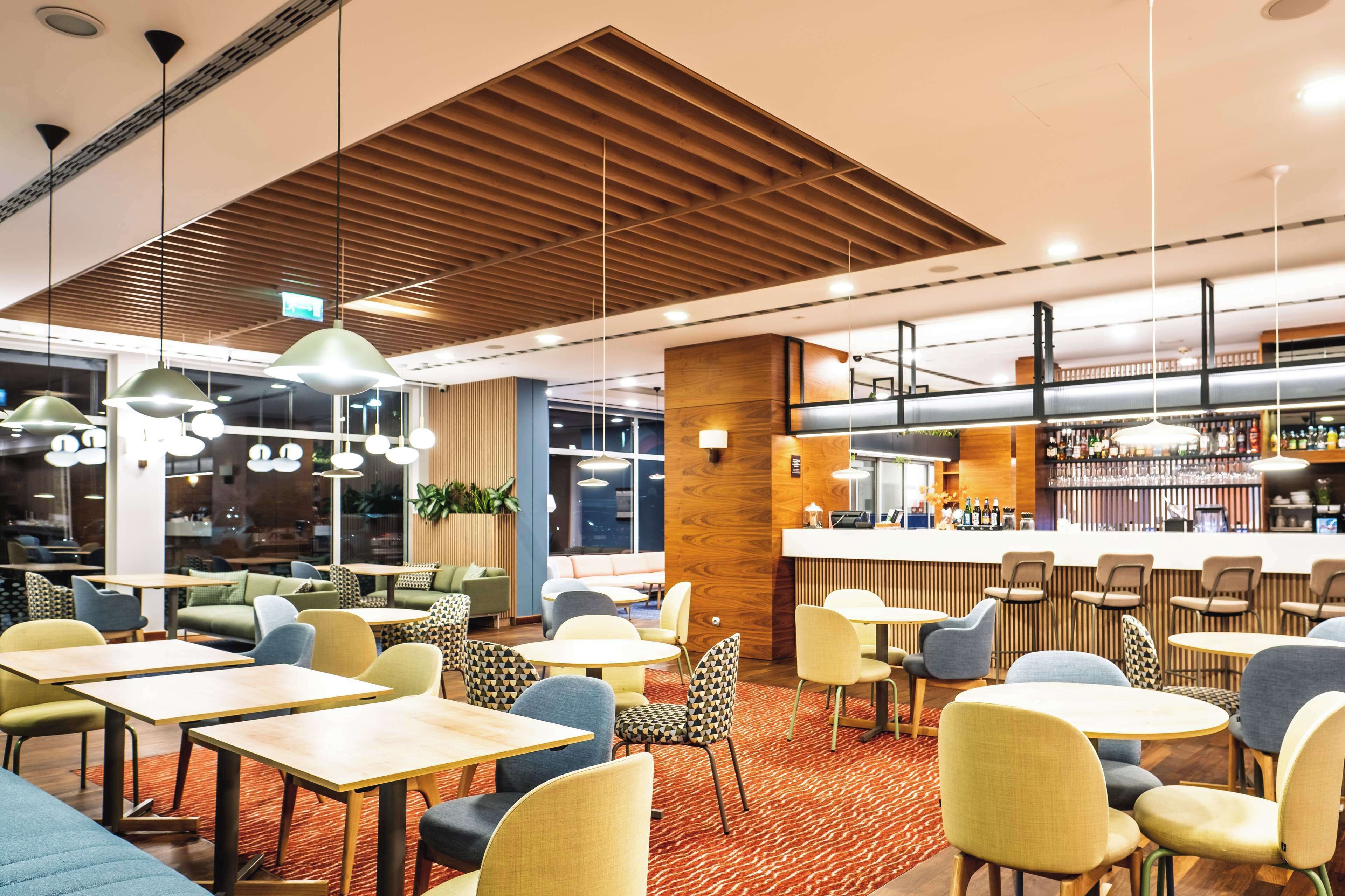 A modern restaurant with wooden ceiling panels, round tables, and colorful chairs.