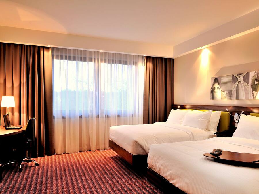 Een hotelkamer met twee bedden, een bureau met een laptop, een stoel en een groot raam met gordijnen.