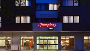 Un hotel llamado Hampton con una fachada moderna, un amplio acceso de vidrio y un área de recepción visible.