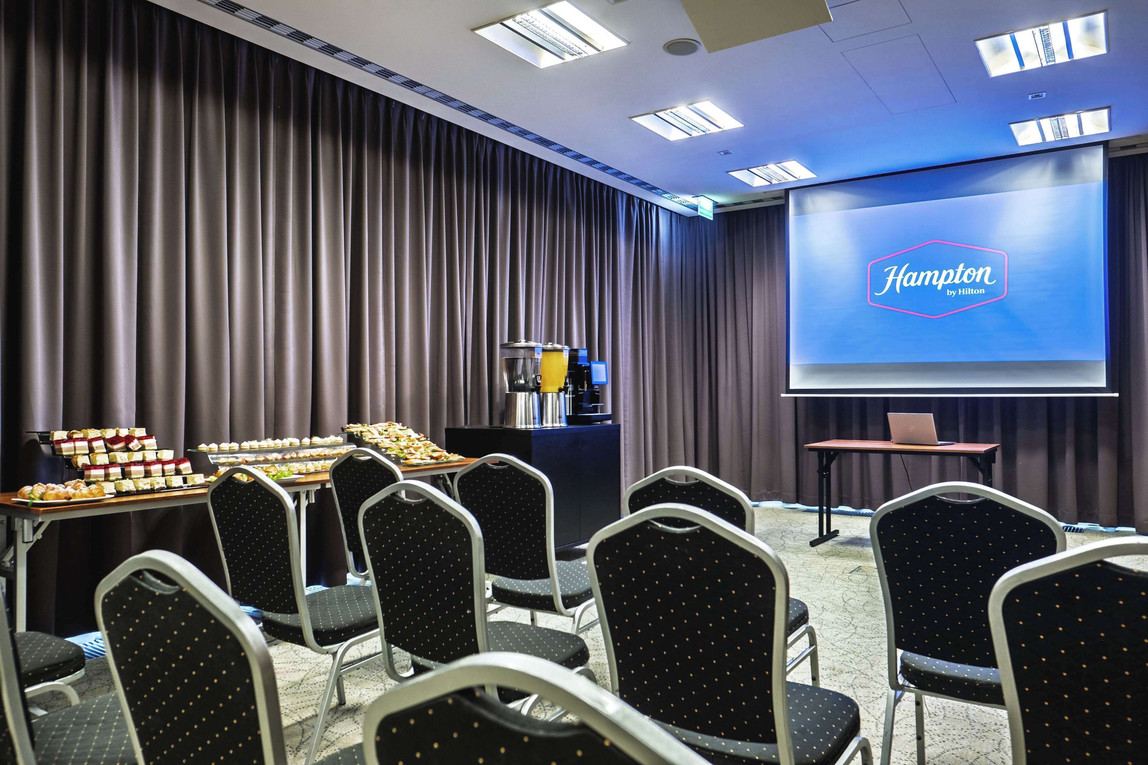 Ett konferensrum med en presentationsskärm som visar texten ”Hampton by Hilton”, ett buffébord med olika sorters mat och rader av stolar.