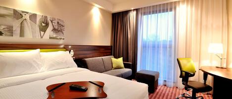 A hotel room with a large bed, a sofa, a desk, and a chair. There is a painting on the wall and a window with curtains.