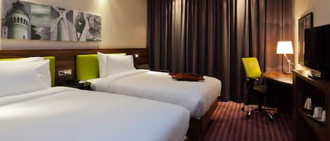 Habitación de hotel con dos camas, un escritorio, una silla, un televisor y obras de arte en la pared.