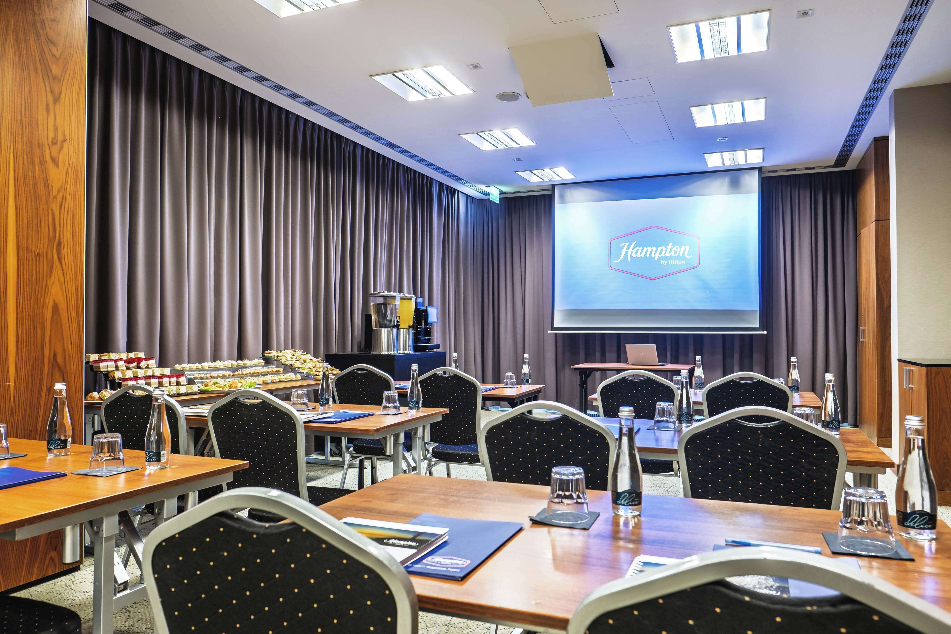 A conference room with a large screen displaying 'Hampton Inn' logo, multiple tables with chairs, water bottles, and a buffet area in the background.