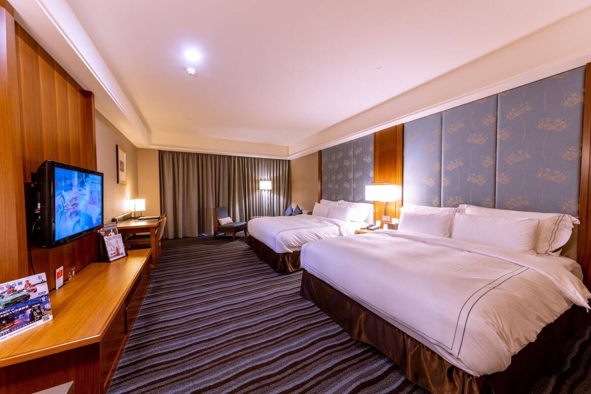 Habitación de hotel con dos camas, televisión, escritorio y un amplio ventanal con cortinas.