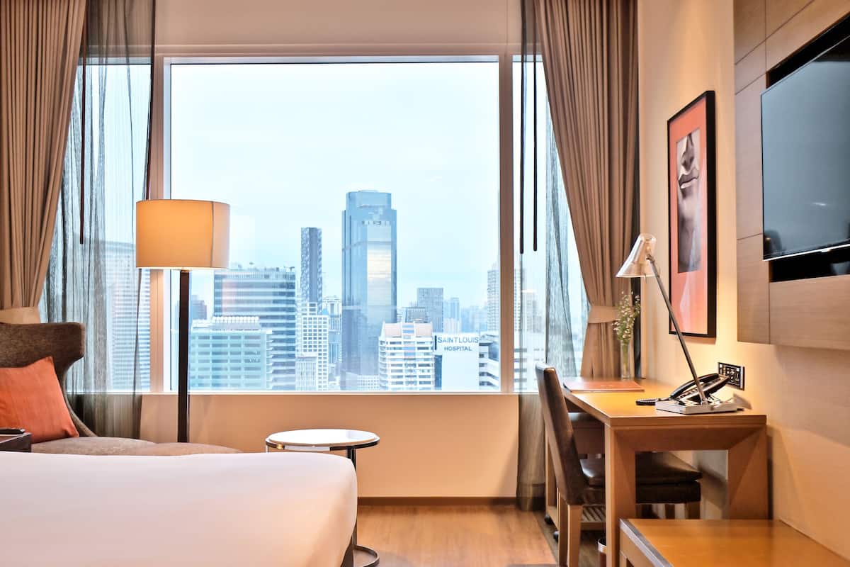Hotelkamer met groot raam en uitzicht over de stad, een bed, bureau met lamp en televisie.