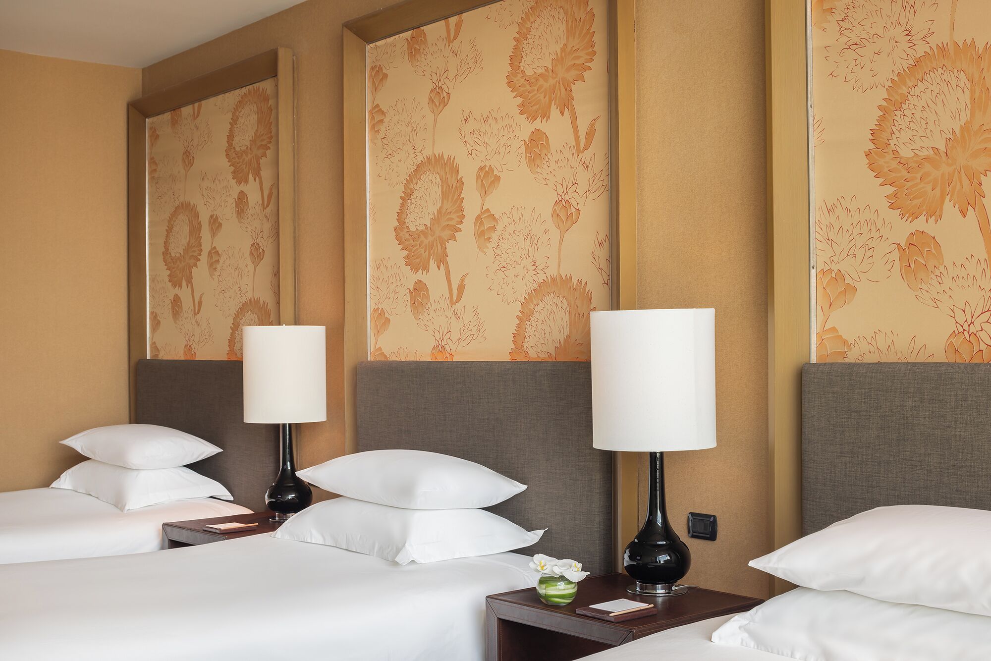 Quarto de hotel com duas camas, cada uma com roupa de cama branca e várias almofadas. Há duas mesas de cabeceira com abajures e um pequeno vaso com flores. O papel de parede das paredes apresenta um padrão floral.