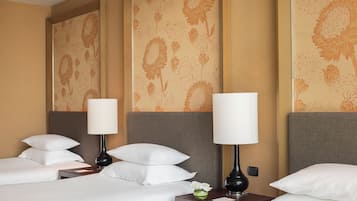 Quarto de hotel com duas camas, cada uma com roupa de cama branca e várias almofadas. Há duas mesas de cabeceira com abajures e um pequeno vaso com flores. O papel de parede das paredes apresenta um padrão floral.
