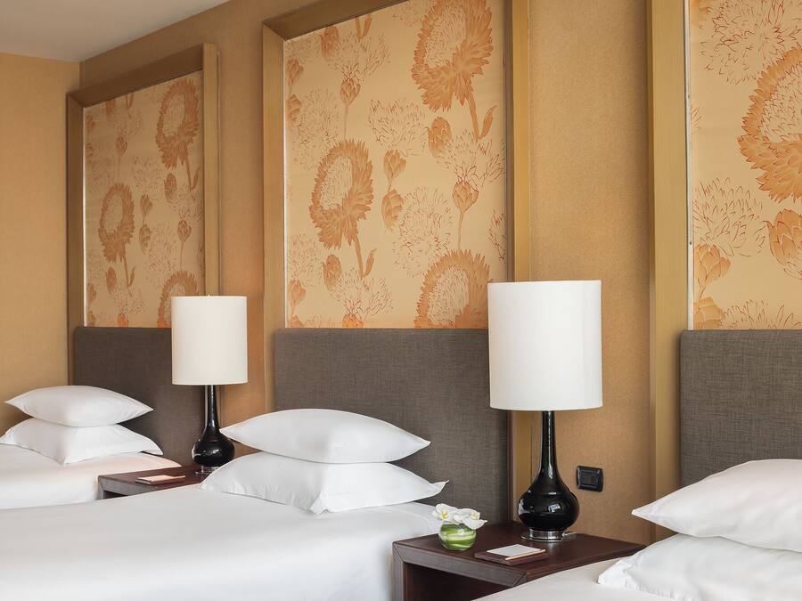 Quarto de hotel com duas camas, cada uma com roupa de cama branca e várias almofadas. Há duas mesas de cabeceira com abajures e um pequeno vaso com flores. O papel de parede das paredes apresenta um padrão floral.