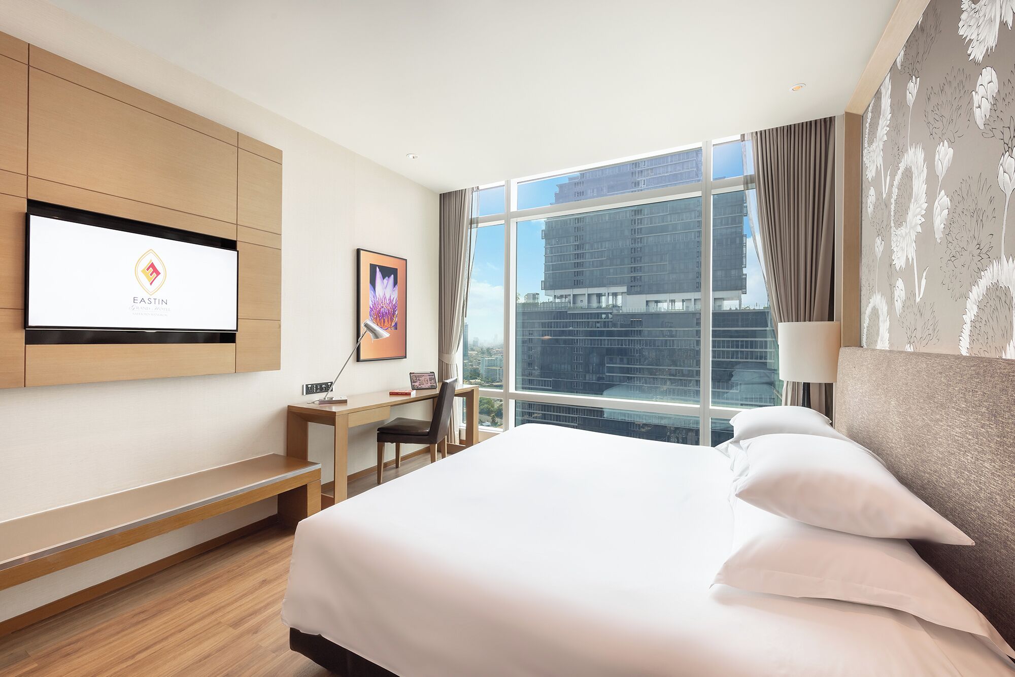 A hotel room with a large bed, a desk, a chair, a TV mounted on the wall, and a large window with a city view.