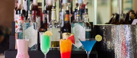 Zona de bar con coloridos cócteles y una selección de botellas de licor al fondo.