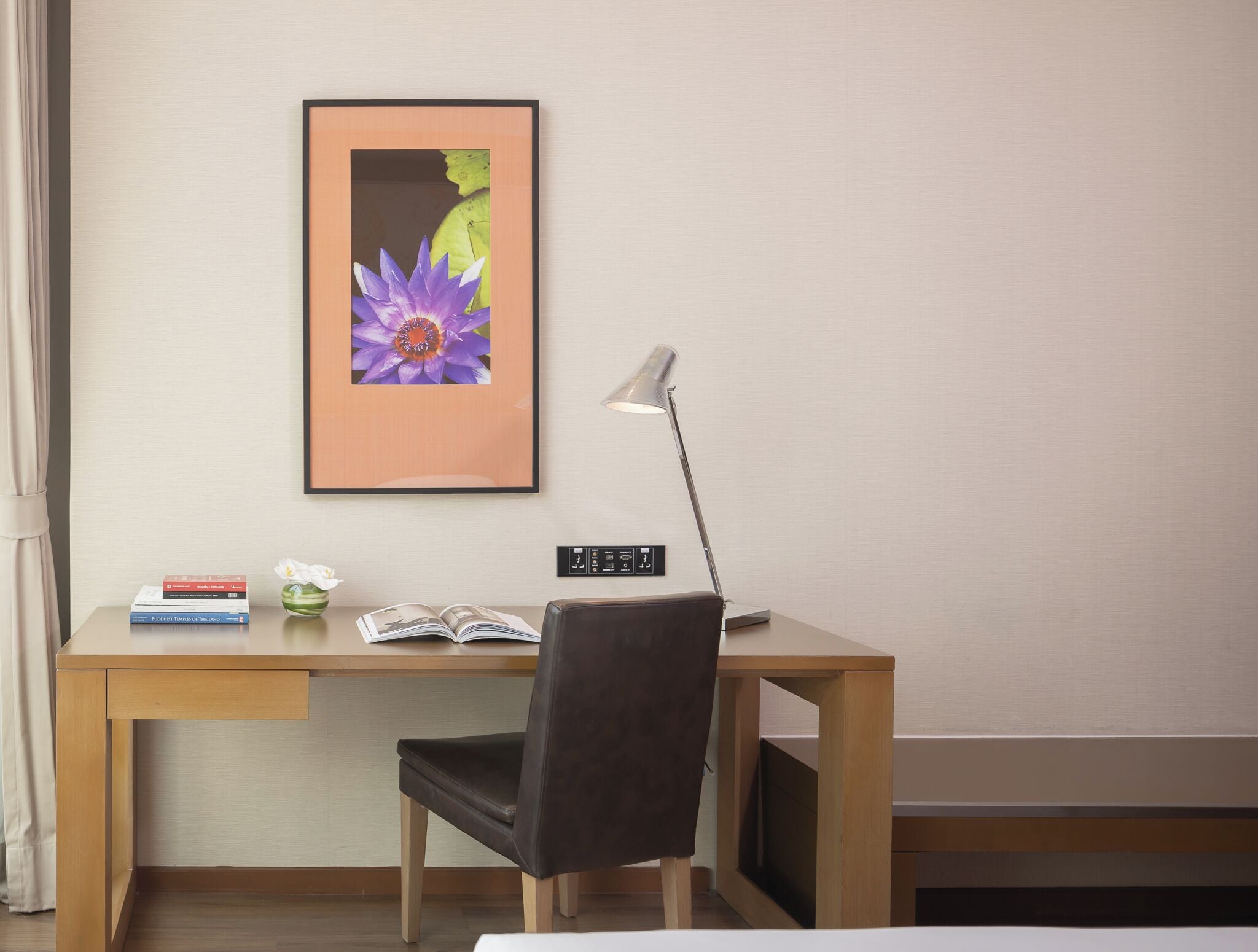 Una habitación de hotel con un escritorio de madera, una silla negra, un cuadro enmarcado de una flor, una lámpara y un libro.