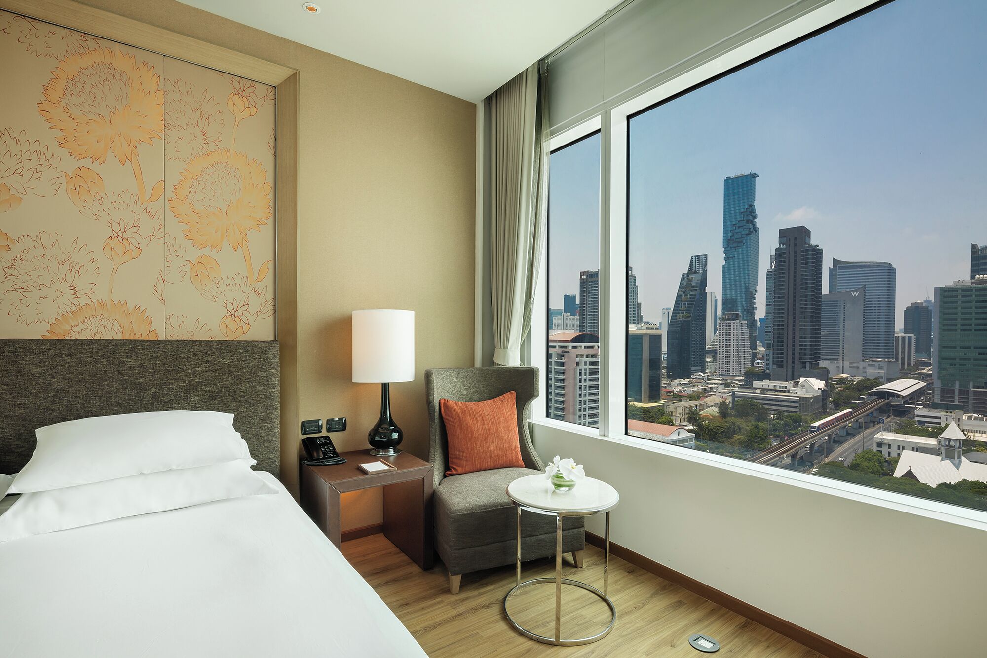Habitación de hotel con cama, una silla, una mesita con un jarrón, una mesita de noche con lámpara y un amplio ventanal con vistas a la ciudad.