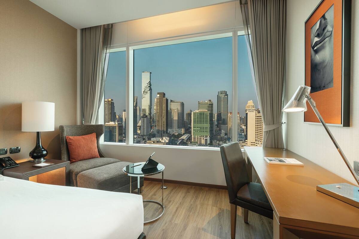 Hotelkamer met groot raam en uitzicht over de stad, een bed, bureau, stoel, lamp en klein tafeltje.