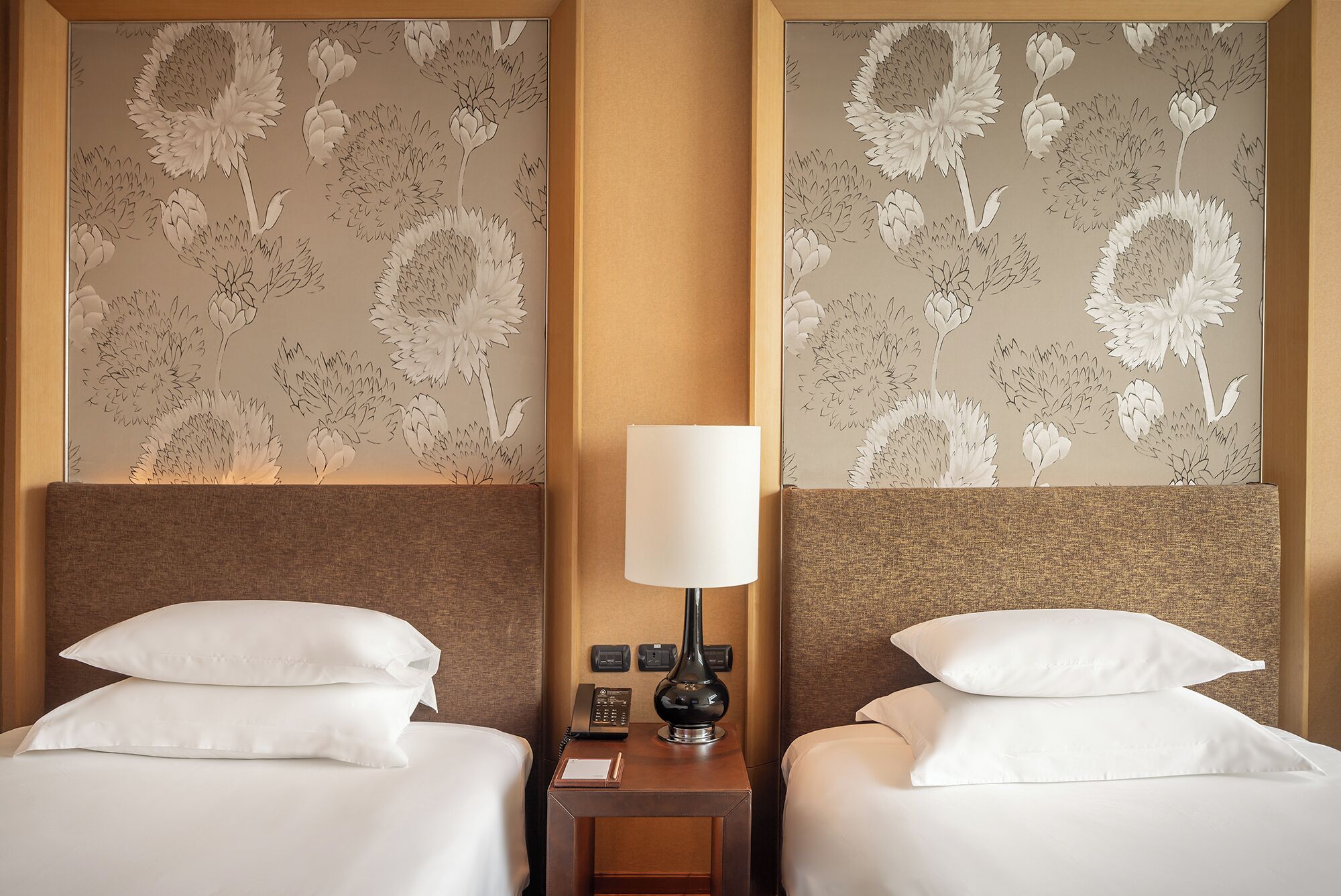 Habitación de hotel con dos camas, una mesita de noche con lámpara y un papel tapiz floral.