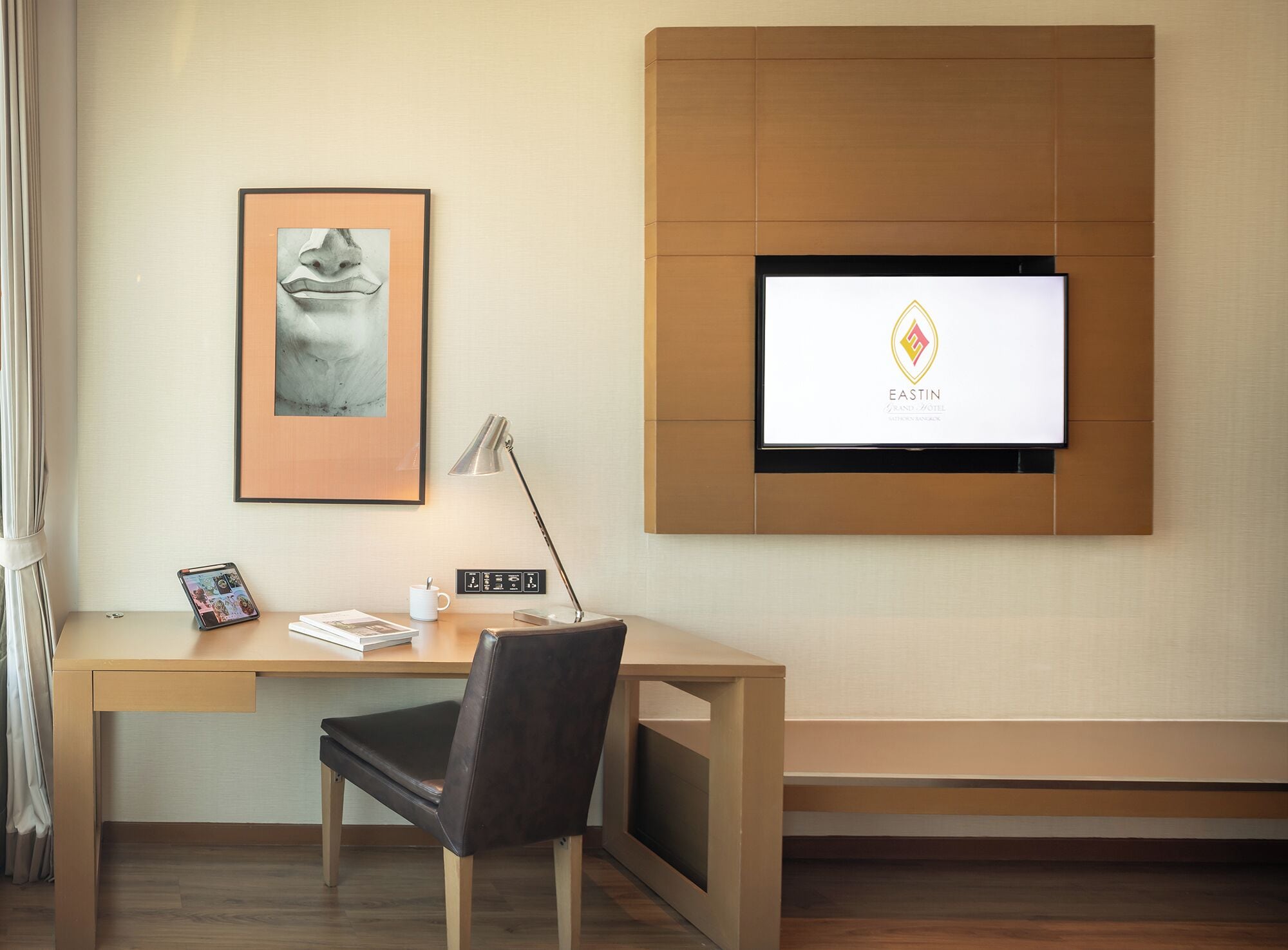 Habitación de hotel con un escritorio, una silla, una obra de arte enmarcada, una lámpara, una taza de café, un teléfono inteligente y un televisor que muestra el logotipo del hotel.