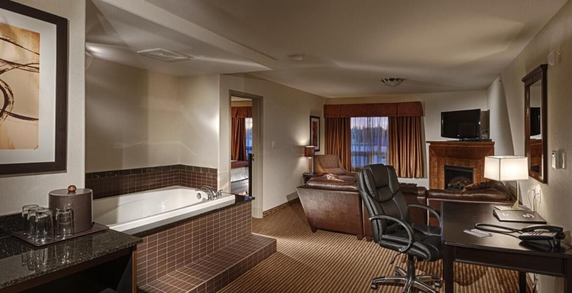 A hotel room with a large bathtub, a desk with a chair, a television, and a fireplace.