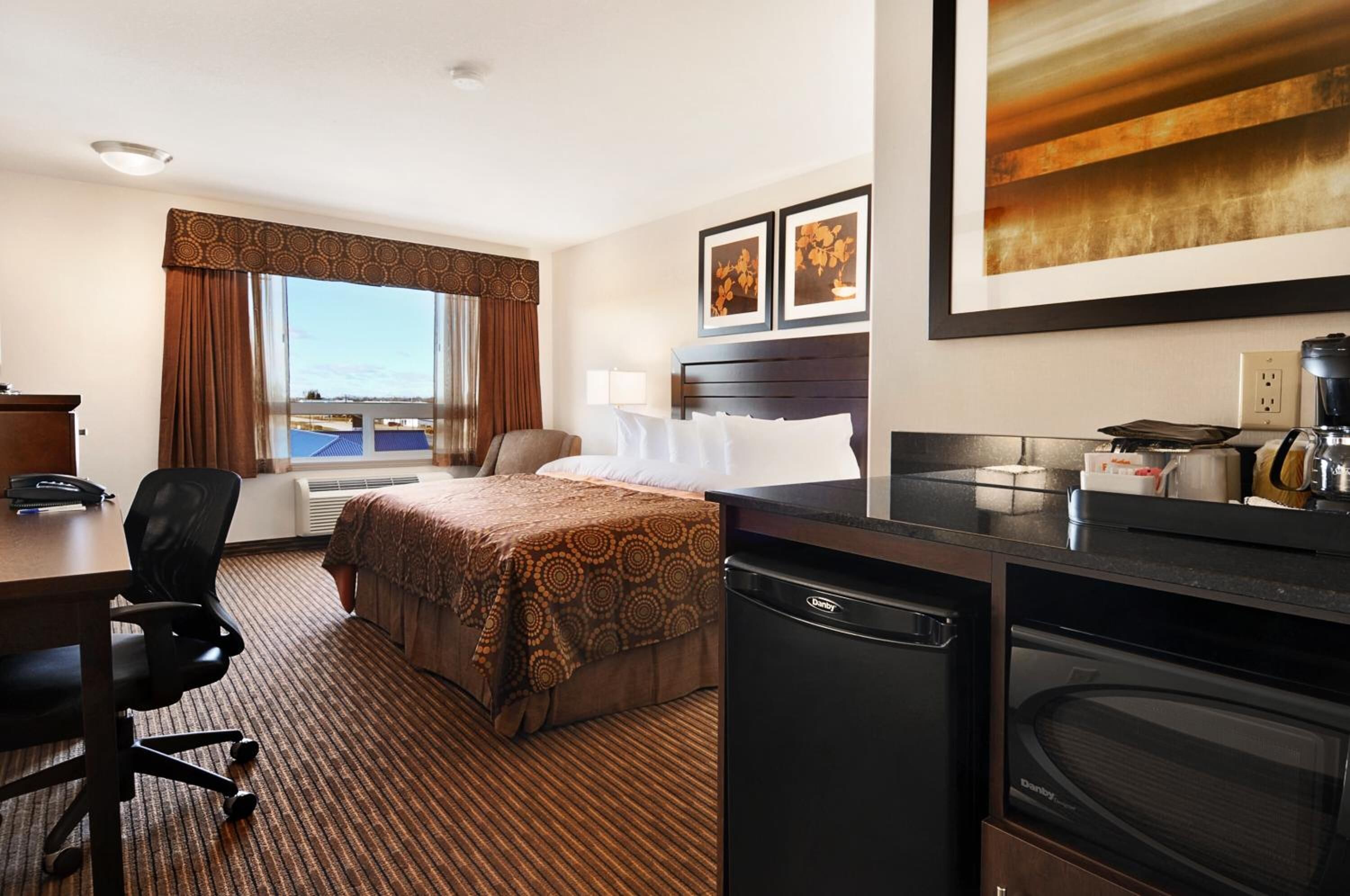 A hotel room with a bed, desk, chair, kitchenette, and a large window with curtains.