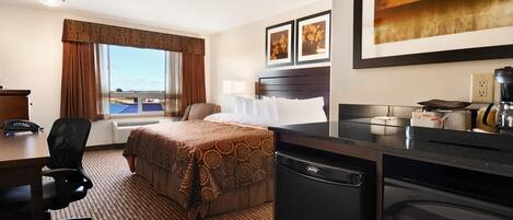 A hotel room with a bed, desk, chair, kitchenette, and a large window with curtains.