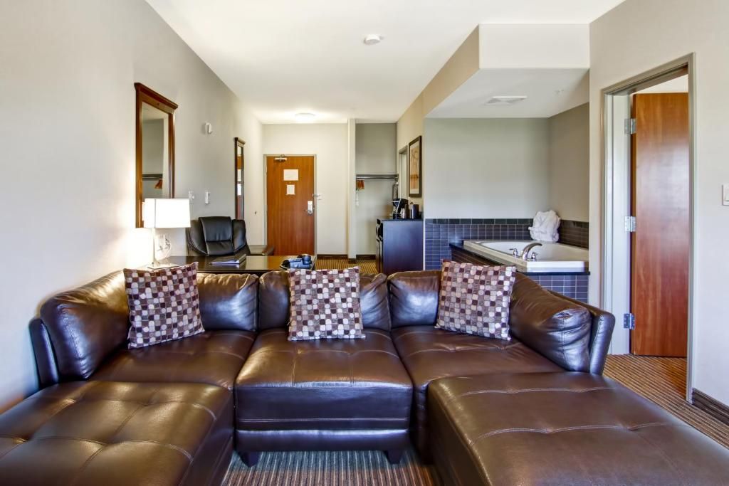 A hotel room with a leather sofa, a desk with a lamp, a chair, a bathroom with a bathtub, and a door leading to another room.
