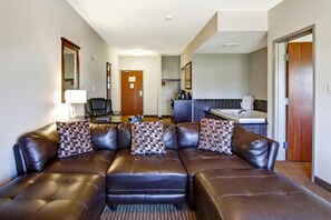 Habitación de hotel con sofá de cuero, un escritorio con lámpara, una silla, un baño con bañera y una puerta que da a otra habitación.