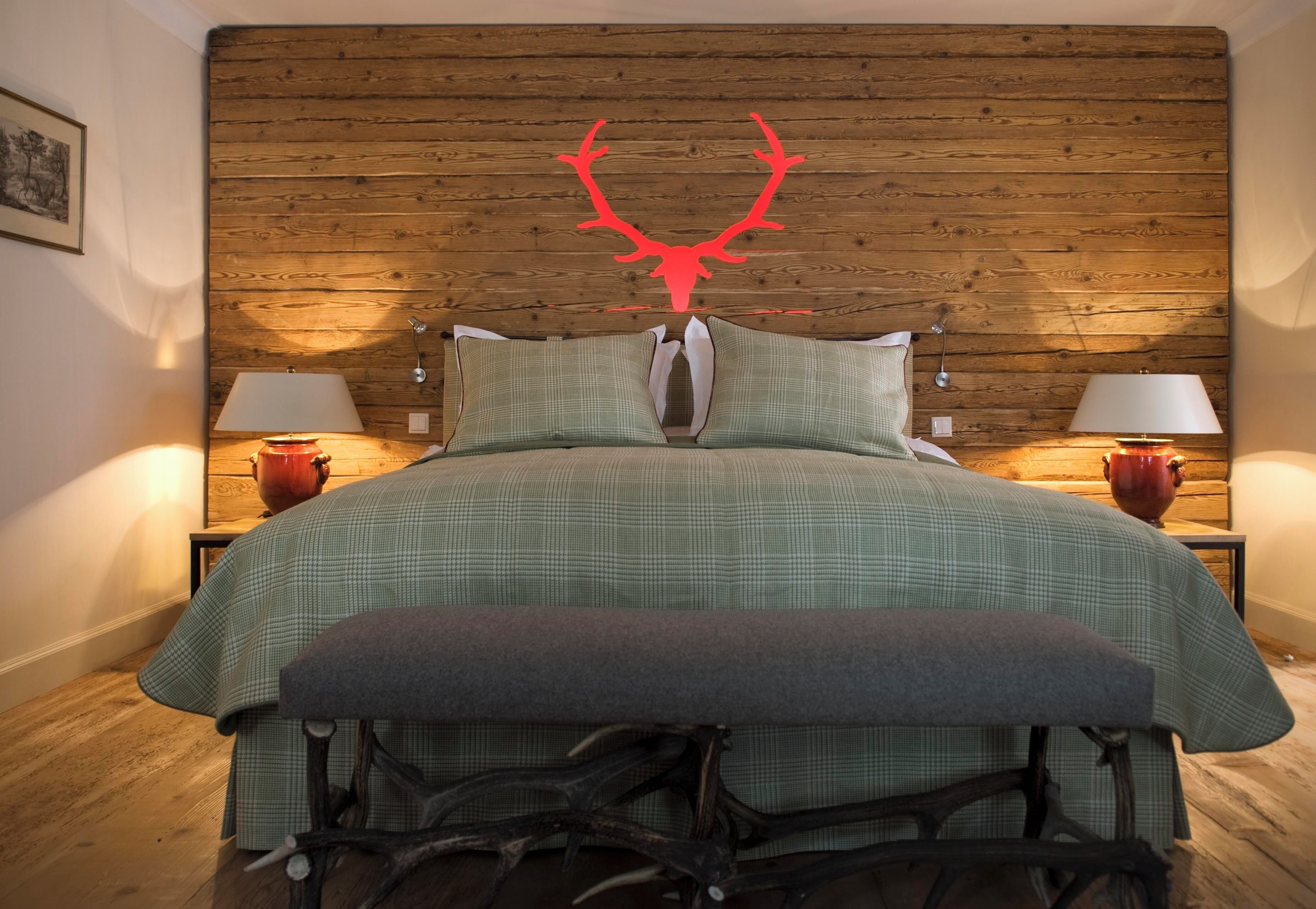 A bedroom with a wooden headboard, two bedside tables with lamps, a green bedspread, and a bench at the foot of the bed.