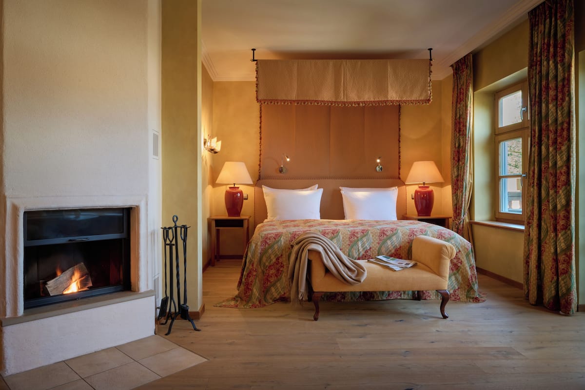 Une chambre à coucher avec une cheminée, un lit recouvert d’une couvre-lit à motifs floraux, deux lampes de chevet et une fenêtre avec des rideaux.