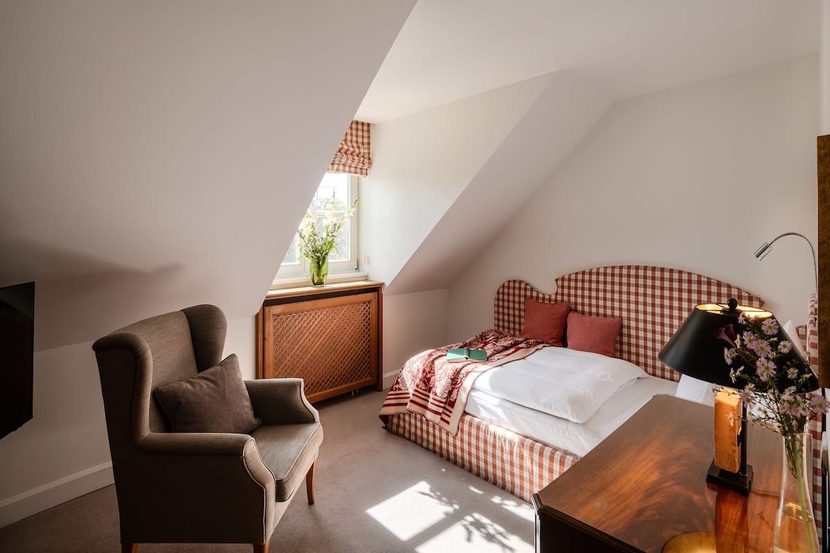 Une chambre avec un plafond en pente, une couvre-lit à carreaux, une table de chevet avec une lampe et une fenêtre donnant sur un paysage verdoyant.