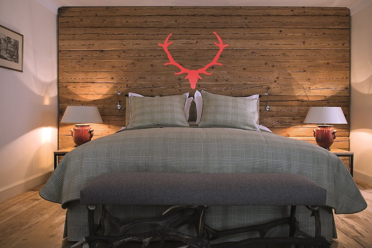 Une chambre à coucher avec une tête de lit en bois, deux tables de chevet avec des lampes, un lit recouvert d’une couverture à carreaux verts et une décoration en forme de bois de cerf rouge.