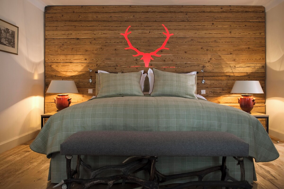 A bedroom with a wooden headboard, two bedside tables with lamps, a green bedspread, and a bench at the foot of the bed.