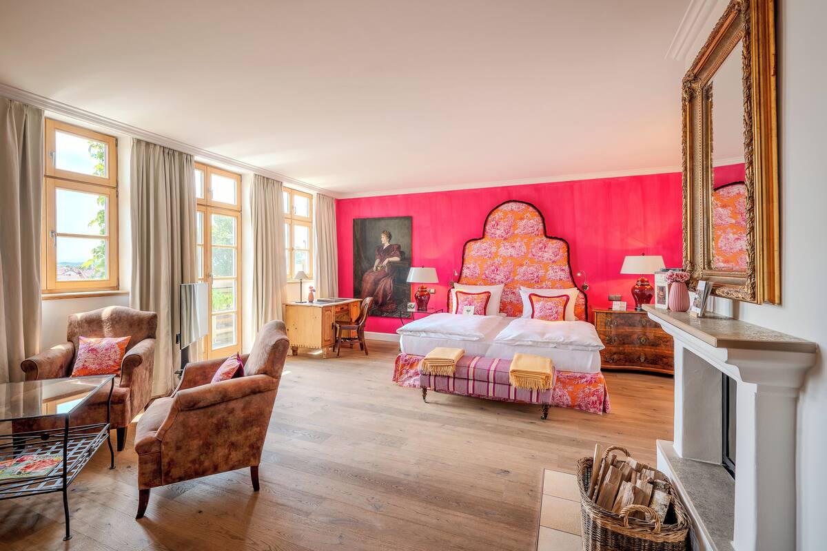 Une chambre avec un grand lit, un bureau et une chaise. On y trouve une cheminée et une fenêtre avec des rideaux.
