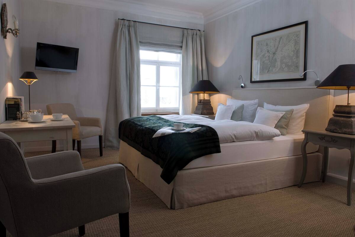 Une chambre d’hôtel comprenant un lit, un bureau avec une chaise, une télévision et une fenêtre avec des rideaux.