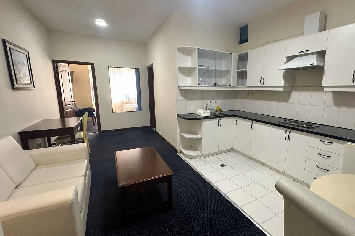 A compact hotel room with a kitchenette, a sofa, a small table, and a window with blinds.