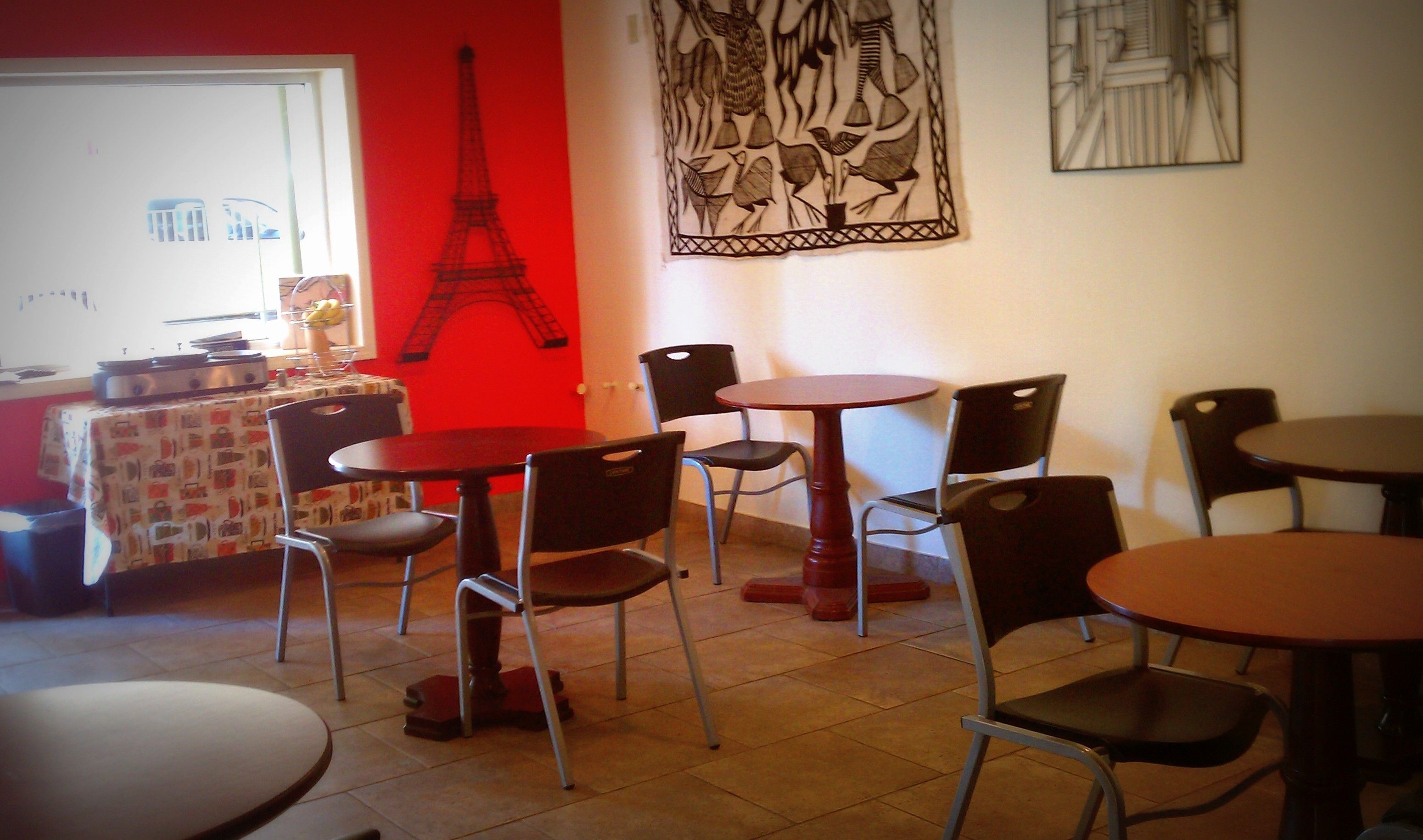 A dining area with multiple tables and chairs, a red wall with a black Eiffel Tower silhouette, and a window with a view of buildings.