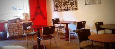 A dining area with multiple tables and chairs, a red wall with a black Eiffel Tower silhouette, and a window with a view of buildings.