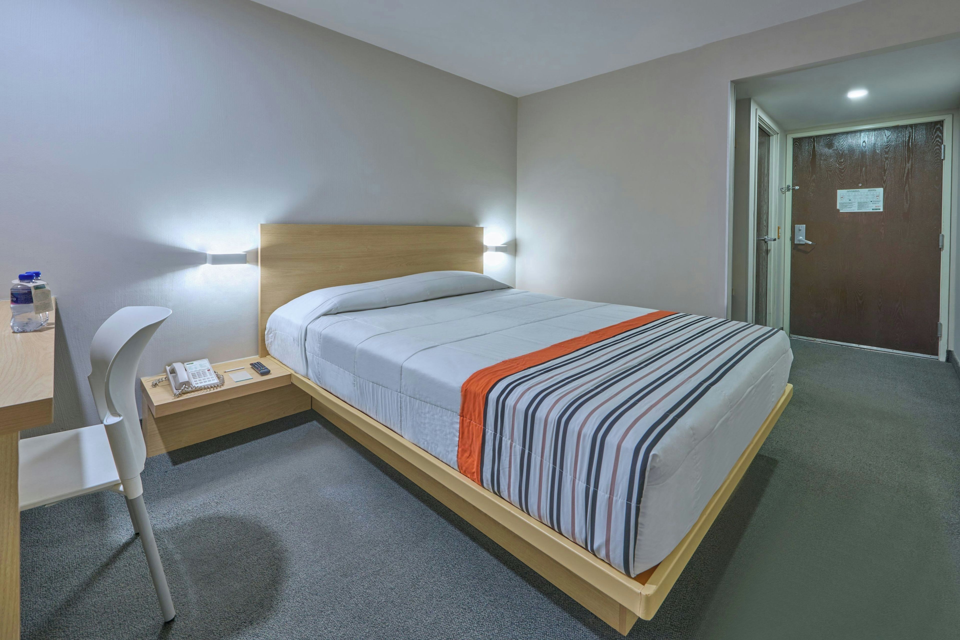 Habitación de hotel con cama, mesita de noche, silla y escritorio con teléfono y botella de agua.