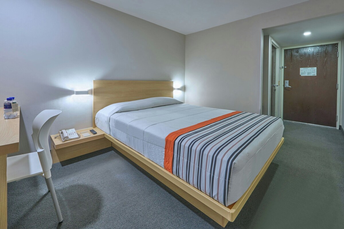 Une chambre d’hôtel comprenant un lit, une table de chevet, une chaise et un bureau avec un téléphone et une bouteille d’eau.