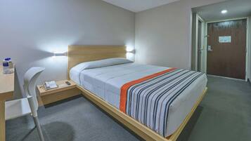 A hotel room with a bed, bedside table, chair, and a desk with a telephone and a water bottle.