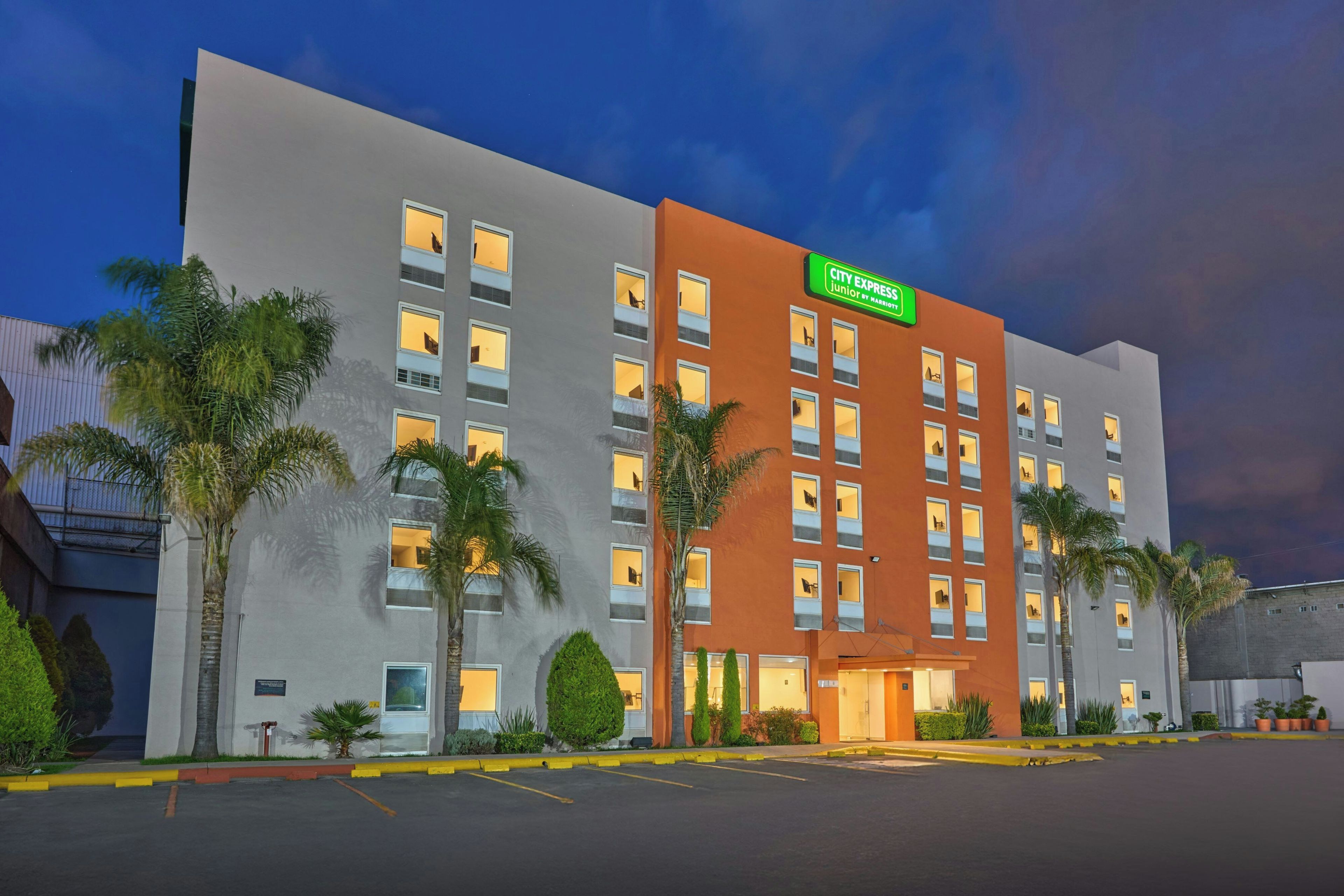 Un hotel de varios pisos con fachada plana, un letrero verde que indica que es un City Express Junior by Marriott, y un estacionamiento al frente.