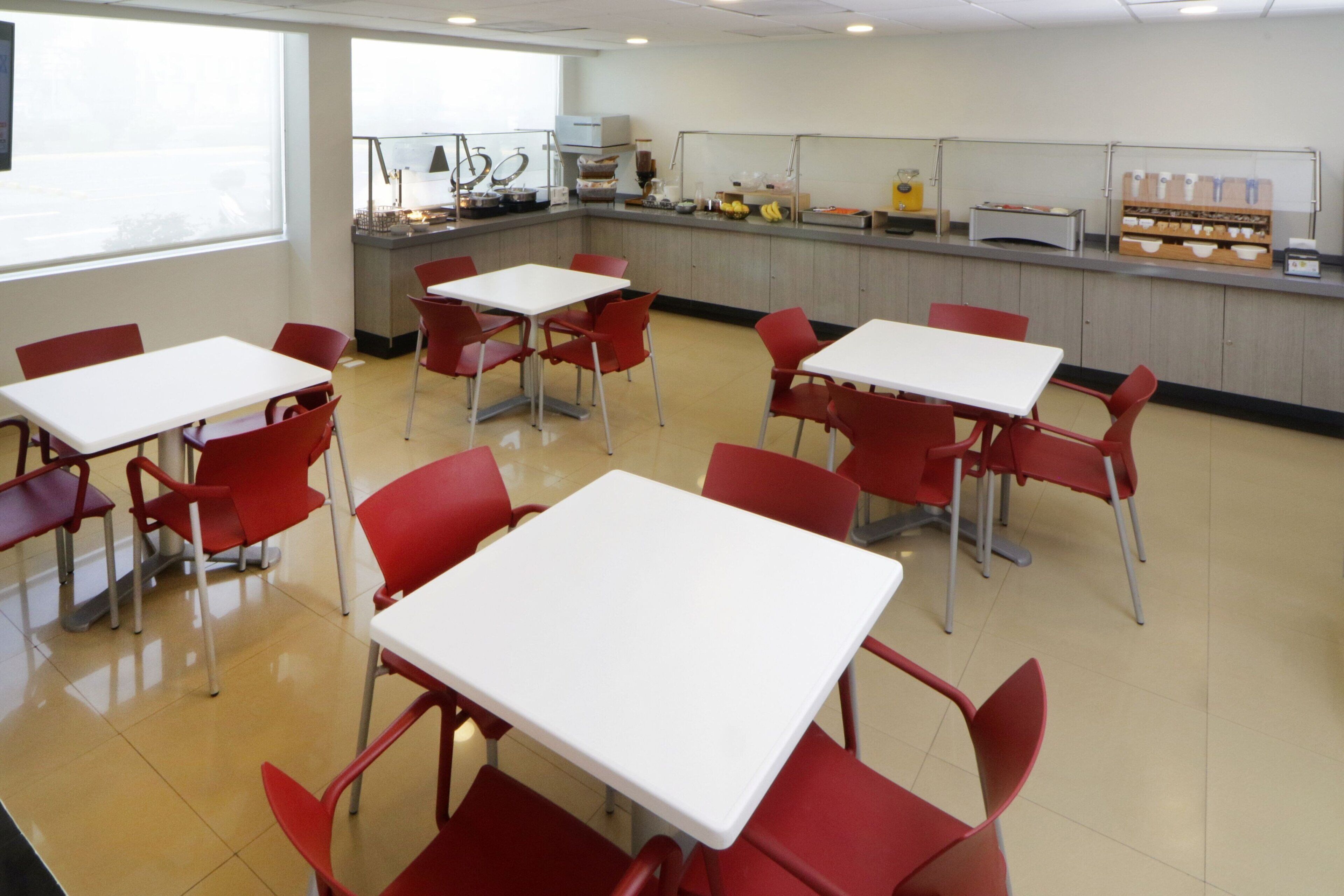 A cafeteria with white tables and red chairs, a counter with food items, and a window providing natural light.