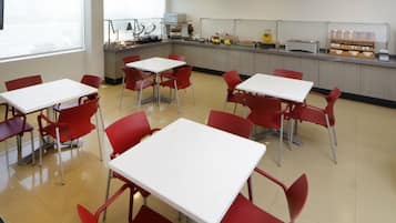 A cafeteria with white tables and red chairs, a counter with food items, and a window providing natural light.