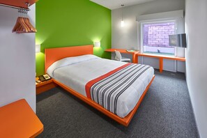 Quarto de hotel com cama, escrivaninha, cadeira, televisão e janela com vista para uma parede de tijolos.