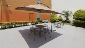 Un patio al aire libre con una mesa y sillas bajo un paraguas grande, rodeado de arbustos bien cuidados y una palmera.