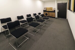 Une salle de conférence avec des rangées de chaises noires, un comptoir en bois sur lequel sont disposés divers objets, et une porte.