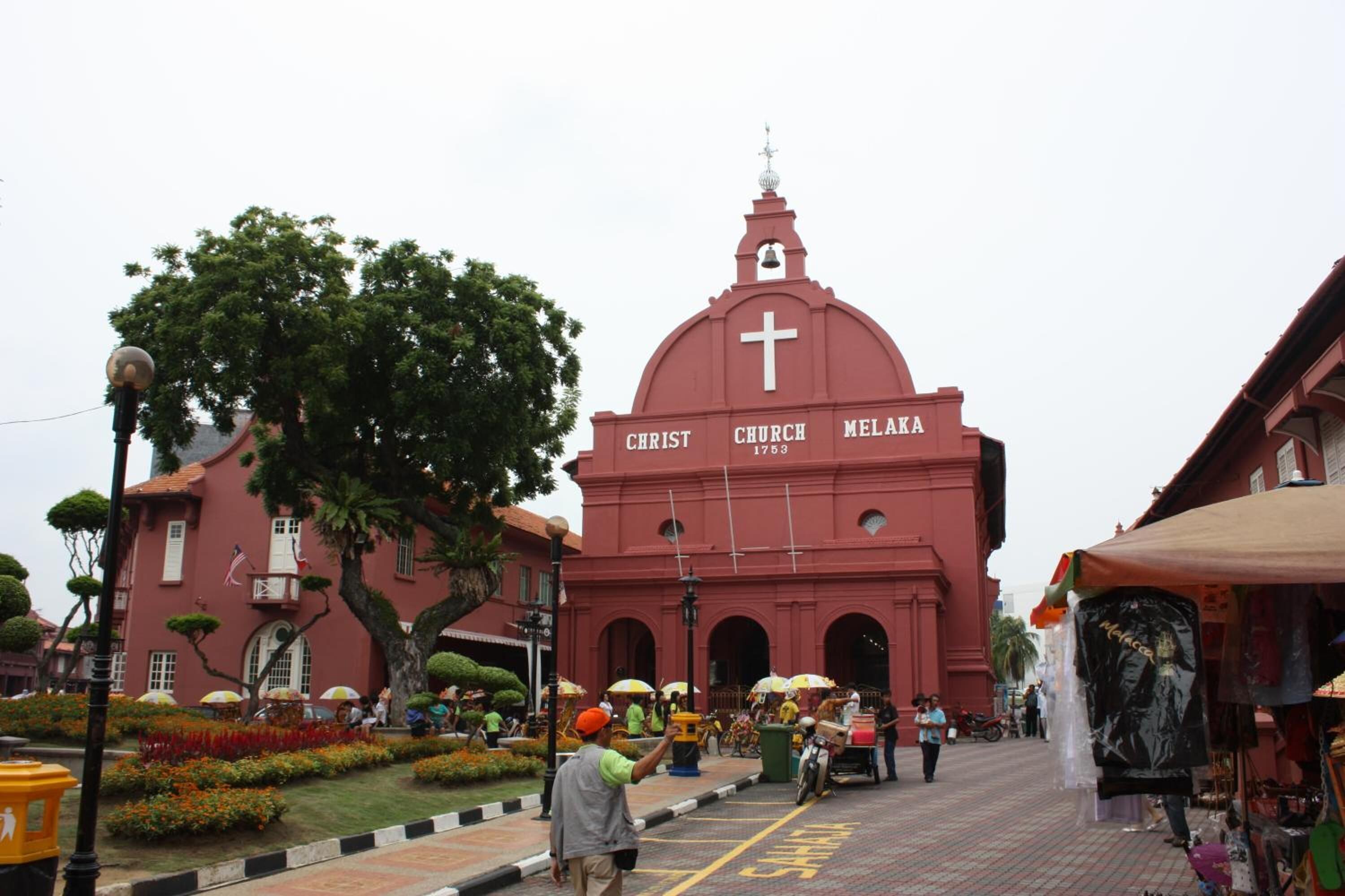 Una iglesia roja con una cruz en la cúpula, rodeada de un jardín cuidado y una calle con puestos de mercado.